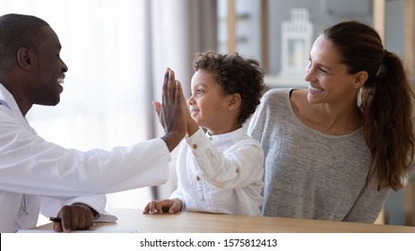 Happy African American Young Male Doctor Giving High Five To Smiling Mixed Race Little Kid Boy, Celebrating Good Result Of Prescribed Medical Treatment Or Welcoming New Patient With Mum At Clinic.