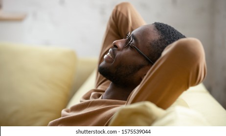 Happy African American Young Man In Glasses Sit Relax On Cozy Couch Lean Hands Over Head Taking Nap In Living Room, Smiling Biracial Male Rest On Sofa At Home Sleeping Or Dreaming, Stress Free Concept