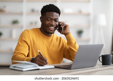 Happy african american young man freelancer or businessman working from home, sitting at table in front of modern laptop, having phone conversation, taking notes and smiling, copy space - Powered by Shutterstock