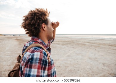 Happy african american young man with backpack standing on the beach and looking far away