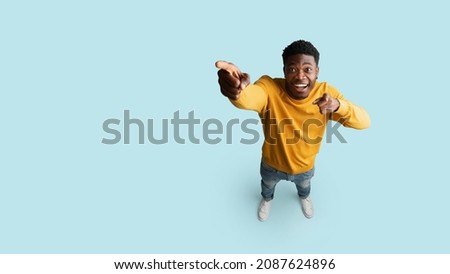 Happy african american young man in casual showing copy space, pointing at blank space for advertisement or text, blue studio background, panorama, top view, full length shot