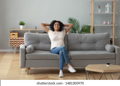 Happy african American young woman sit relax on cozy couch hands over head happy to move to new apartment, smiling black millennial girl rest on comfortable sofa in living room dreaming