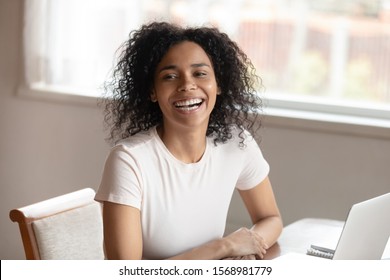 Happy African American Young Woman Sit At Table Work On Laptop Feel Overjoyed Speaking With Someone In Room, Excited Biracial Millennial Female Have Fun Smiling Laughing Communicating Or Talking