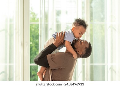 Happy african black parents dad father throw baby son on piggyback and neck riding teasing fun near window. Black baby son and daddy enjoy teasing kiss cheek and neck riding at window light - Powered by Shutterstock