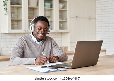 Happy african business man, black male student wearing headphones elearning on laptop computer sitting at kitchen table working from home office, learning online, studying remote training course.