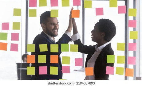 Happy African Business Teacher Woman And Intern Guy Giving High Five, Celebrating Corporate Training Success At Glass Board With Scrum Paper Notes. Diverse Teammates Satisfied With Work Result