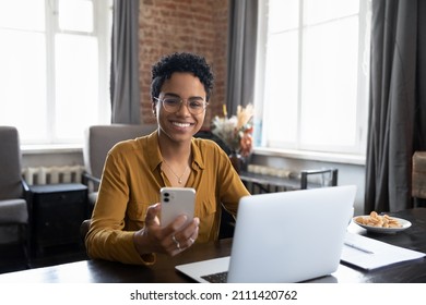 Happy African Business Woman Chatting On Digital Gadgets Online, Holding Mobile Phone, Using Laptop Computer, Looking At Camera, Smiling. Freelance Employee At Home Workplace Head Shot