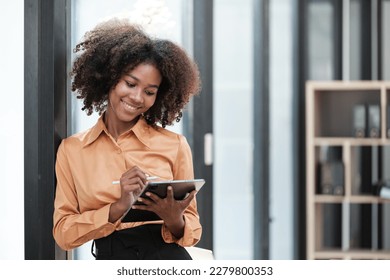 Happy African business woman chatting on digital gadgets online, holding mobile phone, using laptop computer, looking at camera, smiling. Freelance employee at home workplace. - Powered by Shutterstock