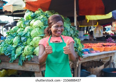 Happy African business woman or female trader wearing a green apron, doing thumbs up gestures while standing at her stall of vegetables in a market place