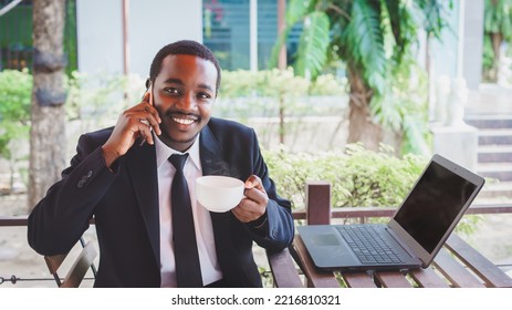 Happy African Businessman In A Suit Use Business Laptop At Coffee Shop. Smiling Black Male Executive Talking By Smartphone And Holding A Cup Of Hot Coffee