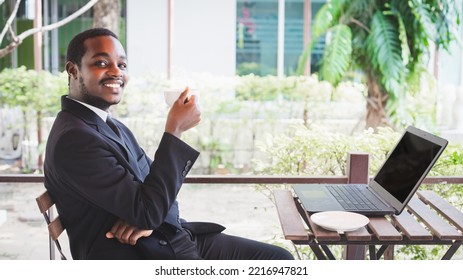 Happy African Businessman In A Suit Use Business Laptop At Coffee Shop. Smiling Black Male Executive Talking By Smartphone And Holding A Cup Of Hot Coffee