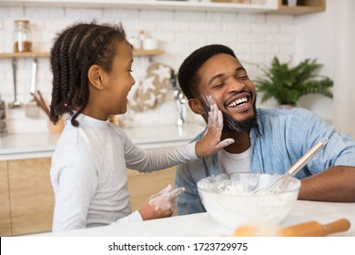 Happy african dad and daughter having fun while cooking pastry at home, girl slapping father face with flour palm