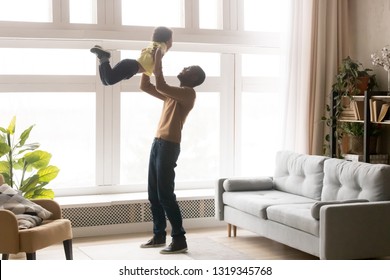 Happy African Dad Lifting Little Toddler Son Up Playing At Home, Loving Black Dad Holding Kid Boy Having Fun In Modern Living Room Together, Small Child Enjoying Weekend Activity Time With Father