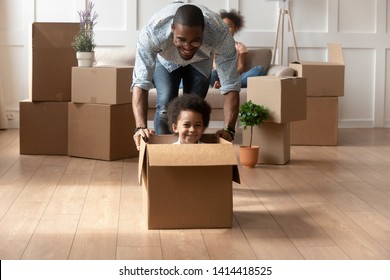 Happy African Dad Playing With Cute Little Mixed Race Son Looking At Camera Laughing On Moving Day, Black Father Having Fun With Kid Boy Riding In Box In Living Room, Relocation, New House Purchase