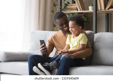 Happy African Dad And Toddler Child Son Laughing Looking At Cellphone Using Funny Smartphone Mobile Face App Sitting On Sofa, Black Father Holding Phone Taking Selfie With Cute Little Kid Boy At Home