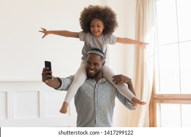 Happy African Dad Wearing Crown Piggybacking Little Funny Daughter Taking Selfie At Home, Loving Black Father Carrying Cute Small Kid On Back Holding Phone Make Photo Playing Together In Living Room