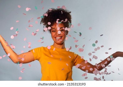 Happy African ethnicity young woman standing with arms outstretched as throwing colored confetti up into air looking with smile while paper petals slowly fall down, posing against blue background - Shutterstock ID 2135313193