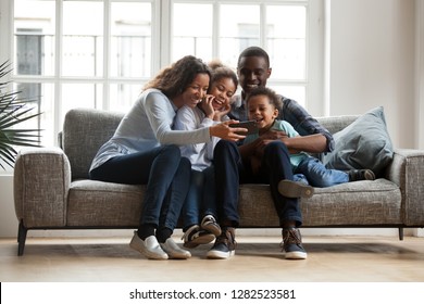 Happy african family with 2 children son daughter having fun with gadget on couch using smartphone at home, black parents and kids laugh watch funny video, make video call selfie on phone online app - Powered by Shutterstock