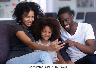 Happy african family with child daughter laugh have fun with gadget on sofa look at smartphone take selfie at home, black parents and kid watch funny video, make online call on phone, use mobile app