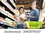 Happy African Family Couple Buying Food In Supermarket, Choosing Products Walking With Cart Along Aisles And Full Shelves Purchasing Groceries Together. Black Spouses Purchasing Essentials Together