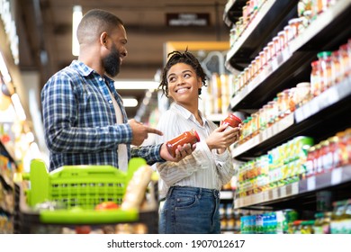 Happy African Family Couple Buying Food In Supermarket, Choosing Products Walking With Cart Along Aisles And Full Shelves Purchasing Groceries Together. Black Spouses Purchasing Essentials Together - Powered by Shutterstock