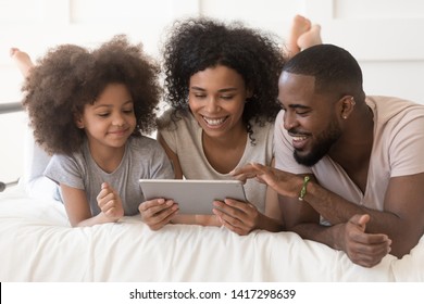 Happy african family with cute little kid daughter using digital tablet lying on bed together, black parents and small child girl having fun looking at computer screen in bedroom in the morning