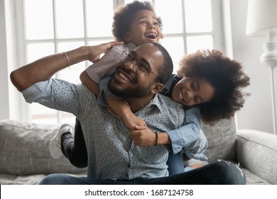 Happy African family at home, cute son and daughter hanging on daddy back, father fool around with little son and daughter piggy back siblings enjoy active time together seated on sofa in living room