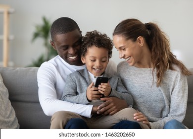 Happy African Family Mixed Race Ethnicity Parents With Small Cute Little Kid Child Son Sit On Sofa Laughing Having Fun Holding Smart Phone Using Funny Mobile App Watching Videos On Cellphone At Home