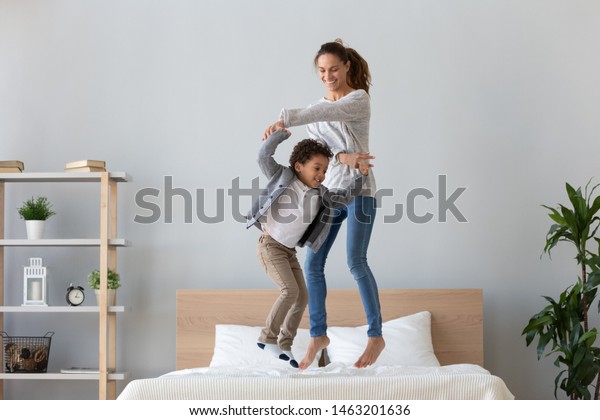 Happy african family mixed race ethnicity kid boy\
and mom baby sitter hold hands jumping on bed mattress, cheerful\
mother having fun laughing playing funny active game with cute\
child son in bedroom