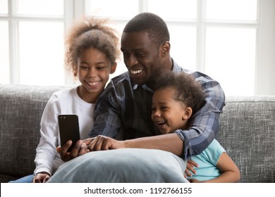 Happy African Family Spending Time Have A Fun Together Using Mobile Phone At Home. Black Father With Little Daughter And Son Make Selfie Photo Picture Watching Video Sitting On Couch At Sitting Room