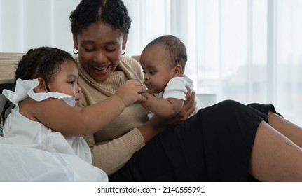 Happy African Family, Young Mother And Daughters, Little Kid Older Sister Looking And Tender Holding Her Newborn Sister Hand While Mom Looking At Children With Love And Care At Bedroom At Home