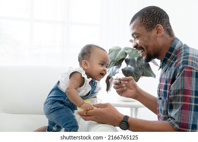 Happy African Father Feeding Adorable Toddler Baby Girl With Spoon While Sitting On Sofa At Home And Kid's Face Mess Up With Food. Little Child Care And Relationship Of Dad And Little Daughter Concept