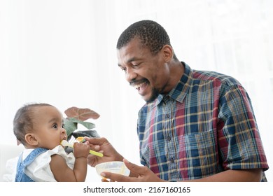 Happy African Father Feeding Adorable Toddler Baby Girl With Spoon While Sitting On Sofa At Home And Kid's Face Mess Up With Food. Little Child Care And Relationship Of Dad And Little Daughter Concept