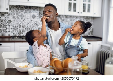 Happy african father holding cute daughters on knees while they feeding him with homemade cookies. Handsome man and two girls having fun during breakfast on modern kitchen. - Powered by Shutterstock