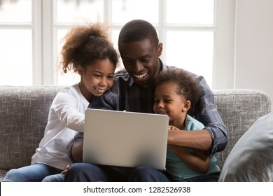 Happy african father and little daughter son using laptop doing family online shopping on sofa, black dad watching funny video with kids on computer, daddy and children having fun playing with app