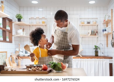 Happy African Father And Son Dress Up Together Before Cooking In The White Kitchen. Single Dad Chef With Black Kid Helper In Yellow Shirt Preparing Food At Home. Eye Contact, Relationship, Warm Family