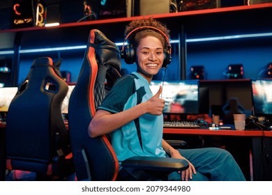 Happy African female cybersport gamer wearing headset showing approval sign while sitting on gaming chair in internet cafe