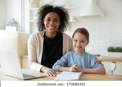 Happy African Female Tutor And Little Learner Schoolgirl Portrait. Multi-racial Family Sit In Kitchen Laptop And Workbooks Lie On Table. Successful Homeschooling, Clever Kid Girl Study At Home Concept