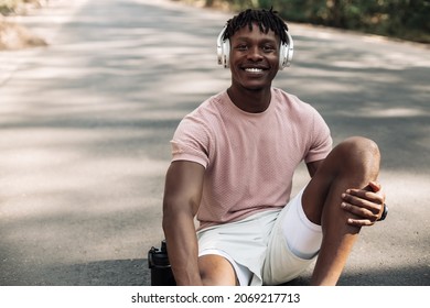 Happy African Fitness Man Sitting Relaxed On The Road In The Park, With A Bottle Of Water After Workout, Sportsman Taking A Break After Exercise Outdoors In Nature