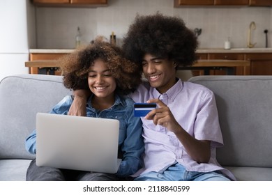 Happy African Gen Z Teen Student Couple Hugging On Couch, Enjoying Online Shopping From Home, Paying By Credit Card, Making Payments For Purchase From Virtual Account, Using Laptop Computer