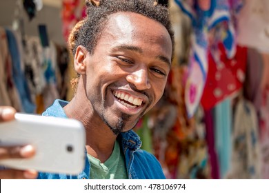 Happy African Guy Is Spontaneous, Smiling And Taking A Selfie Photo Using Smartphone Out In A Colorful Market Place - Cheerful Man Looking At The Phone With A Wide Toothy Smile On A Bright Sunny Day