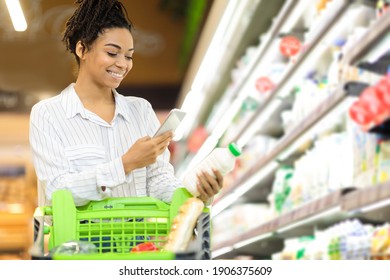 Happy African Lady Scanning Product On Grocery Shopping Buying Food In Supermarket. Female Buyer Using Phone App Choosing Dairy Products Standing With Shop Cart In Store. Free Space