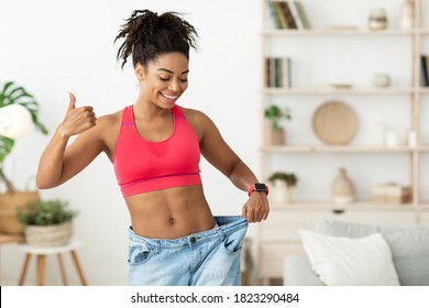 Happy African Lady Showing Result Of Successful Weight Loss Gesturing Thumbs-Up, Wearing Old Oversize Jeans Standing At Home. Slimming Motivation And Dieting Concept. Blank Space For Text