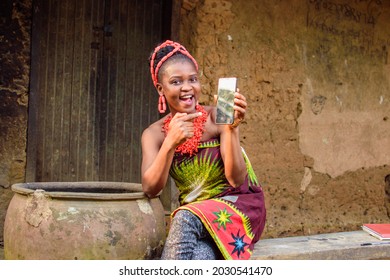 A happy African lady or woman with beads on her head, sitting beside big water calabash, excitedly pointing to a smart phone outside a village mud house