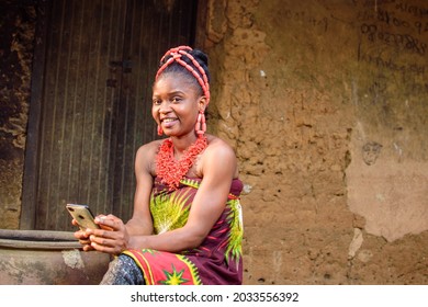 A happy African lady or woman with beads on her head, sitting beside big water calabash, excitedly holding a smart phone outside a village mud house