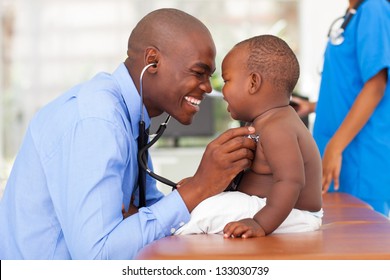 Happy African Male Doctor Examining Baby Boy With Female Nurse On Background