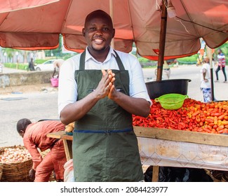 A happy African male trader with an apron standing beside his stall of tomatoes and pepper in a market