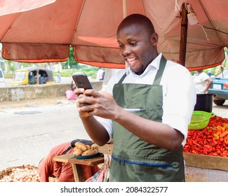 A Happy African Male Trader With A Smart Phone, Standing Beside His Stall Of Tomatoes And Pepper In A Market Place