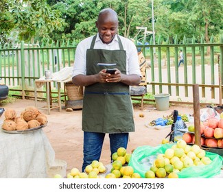 Happy African male trader or vendor holding a smart phone while standing at his fruit stall in a market place