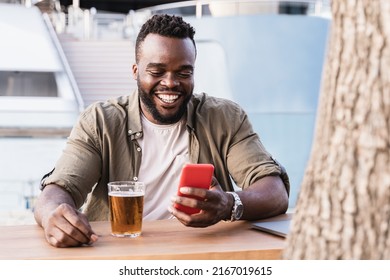 Happy African man drinking beer at brewery bar using mobile phone outdoor with luxury yacht in background - Focus on face - Powered by Shutterstock